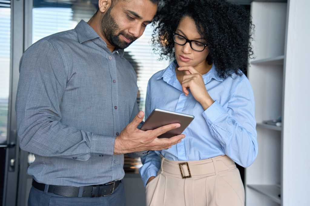 Focused Indian male IT leader and female African American financial business leader discussing financial business project looking at tablet device standing in modern corporate office.