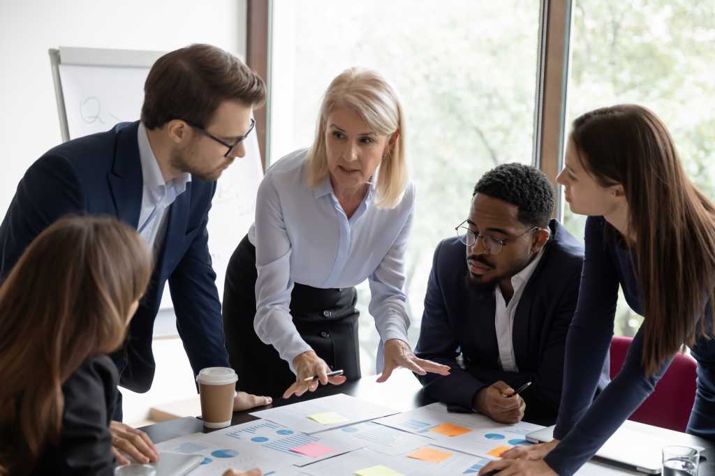 Mature business teacher training group of employees, teaching to work with paper reports, analyze marketing charts, using sticky notes. Mentor explaining scrum method tools to employees