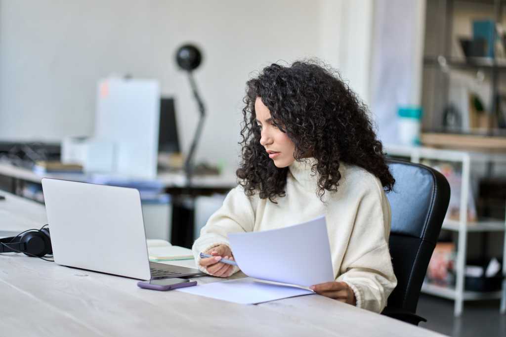 Young latin business woman manager accounting analyst checking bills, analyzing sales statistics management, taxes financial data documents or marketing report papers working in office using laptop.